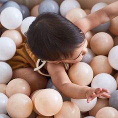 a baby in a ball pit with lots of balloons on the floor and one hand reaching for it