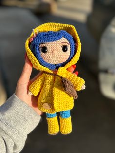 a hand holding a small crocheted doll wearing a yellow jacket and blue pants