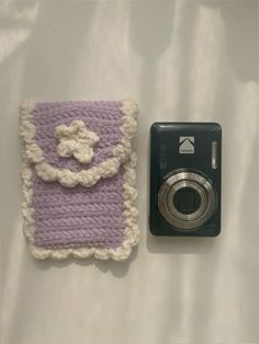 a camera next to a crocheted case on a white sheet with a purple and white flower