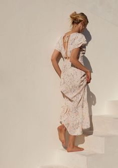 [Color: Natural/Peach] A back facing image of a blonde model standing outside wearing a bohemian special occasion maxi dress in a natural and pink floral print. With short flutter sleeves Bohemian Ditsy Floral Maxi Dress For Brunch, Feminine Boho Print Maxi Dress, Beach Midi Dress With Flutter Sleeve And Smocked Back, Flutter Sleeve Midi Dress With Smocked Back For Beach, Feminine Floral Dress With Flutter Sleeves For Beach, Feminine Maxi Dress With Smocked Back And Flutter Sleeves, Feminine Flutter Sleeve Maxi Dress With Smocked Back, Summer Peach Floral Print Maxi Dress, Bohemian Midi Dress With Flutter Sleeves And Floral Print