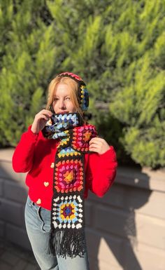 a woman wearing a red sweater is holding a cell phone up to her ear and has a crocheted scarf around her neck