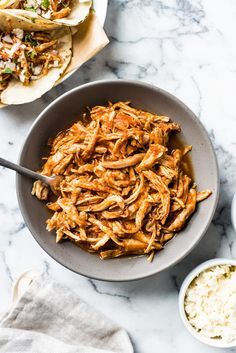 a bowl filled with shredded chicken next to two tortillas on a marble counter