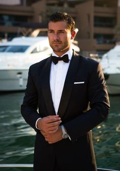 a man in a tuxedo standing next to some water with boats behind him