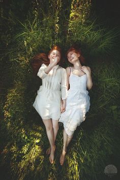 two women laying in the grass with their arms around each other, both wearing white dresses