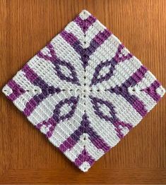 a crocheted square with purple and white designs on it sitting on top of a wooden table