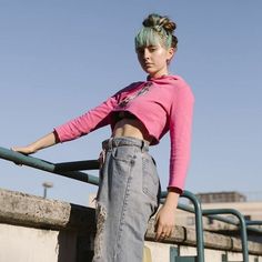 a woman with blue hair and pink shirt leaning against a metal rail on a sunny day