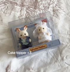 two small stuffed animals are sitting in a plastic box on a white bed sheet with the caption cake topper = 3