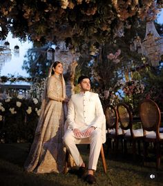 a bride and groom sitting on chairs under a tree with flowers hanging from the branches