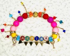 "\"ARRIBA\" Atinys!! Rep the most fun track of their blockbuster WILL album! If you love color, this one's for you! Vibrant hues of pink, yellow, orange and blue paired with bright gold trim with rhinestone and pavé accents, embellished with bright faceted glass and gold flag hanging embellishments, it's a fiesta on your wrist! Adorned with a gold filled margarita glass charm and whimsical music note charm with micro pavé stones. Letters are metal alloy with multicolor enamel. Sizes are approximate based on bead size, can vary within 1/8-1/4\"." Novelty Multicolor Bracelets For Party, Multicolor Novelty Bracelets For Party, Fun Multicolor Beaded Bracelets For Party, Personalized Multicolor Beaded Bracelets For Party, Fun Pink Beaded Bracelets For Party, Flag Hanging, Music Note, Bright Gold, Faceted Glass