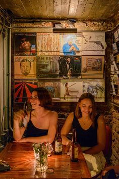 two women sitting at a wooden table with bottles and glasses in front of the wall