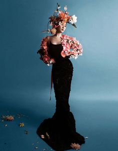 a woman in a black dress with flowers on her head is standing against a blue background