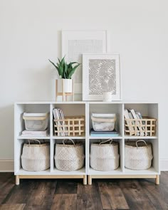 a white shelf with baskets and flowers in it on top of a wooden floor next to a wall