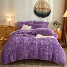 a bed with purple comforter and pillows in a room next to a lamp on a table