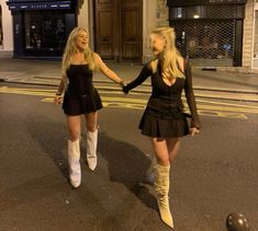 two women dressed in short skirts and boots walking down the street holding each other's hands