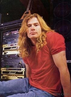 a man with long hair sitting in front of a rack of musical equipment and looking at the camera