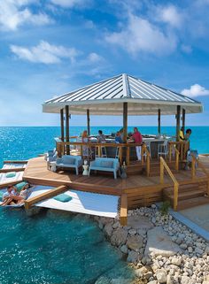 people are sitting at tables on the dock by the ocean with an umbrella over them