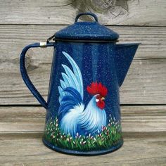 a painted blue coffee pot with a rooster on it's side and grass in the background