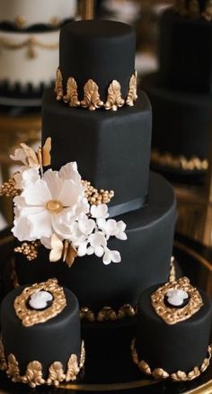 a black and gold wedding cake with white flowers on the top tier is surrounded by other cakes