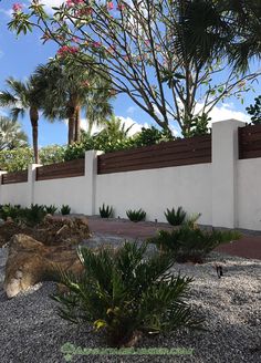 an outdoor area with rocks and plants next to a white wall that has a wooden fence on it