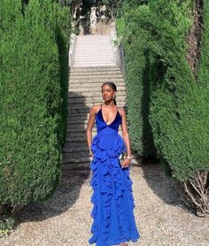 a woman in a blue dress standing next to some bushes and stairs with her hands on her hips