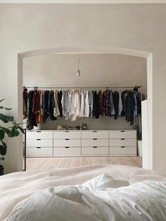 a bed sitting under a window next to a closet filled with shirts and pants on hangers