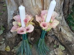 two white candles with pink flowers and green leaves on them next to a tree trunk