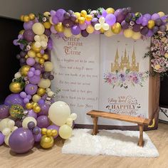 a purple and gold balloon arch with a wooden bench in front of it, surrounded by balloons