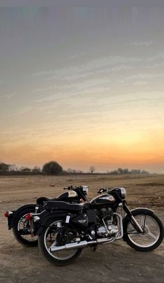 two motorcycles are parked in the dirt near each other on an empty field at sunset