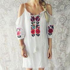 Gorgeous Mexican embroidered details adorn this bohemian tunic dress. Designed with a white base, off-the-shoulder sleeves, front tassel tie, and embroidered details throughout. Perfect paired with sandals for a breezy summer outfit. One size for small, medium, & large. Custom made to order. Please allow 2 weeks for production. Color: White & multi 100% Cotton Imported Hand Wash Cold One size for S,M, & L Bust 37 Waist 38 Hips 39 Length 35 Bust, waist, and hip measurements are a tota Mexican Boho, Bohemian Tunics, Mexican Fashion, White Boho Dress, Bohemian Bags, Embroidered Details, Wholesale Dress, Boho Women, Dress First