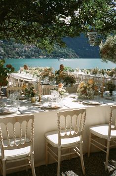 an outdoor table set up with white chairs and flowers on it, overlooking the water