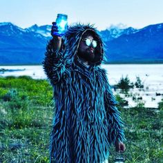 a man in a blue furry coat holding a water bottle and wearing sunglasses with mountains in the background