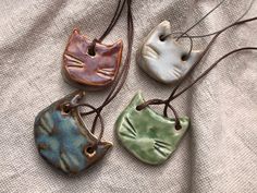 three ceramic cats are hanging from leather cord necklaces on a white cloth background,