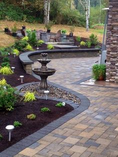 a fountain in the middle of a brick patio surrounded by trees and shrubs, with potted plants on either side