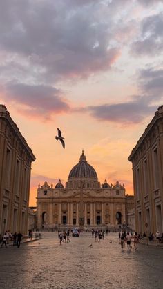 people are walking around in front of a building with a bird flying over it at sunset