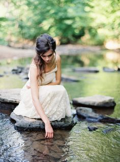 a woman in white dress sitting on rocks by water with trees in the back ground