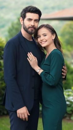 a man and woman standing next to each other in front of some bushes with trees