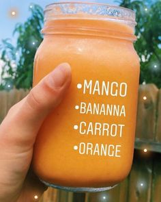 a hand holding up a jar of orange smoothie with the words mango, banana, carrot and orange on it