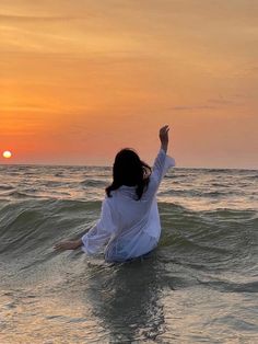 a woman sitting on top of a surfboard in the ocean at sunset with her arms up