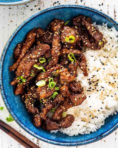 a blue bowl filled with meat and rice next to chopsticks on a white table