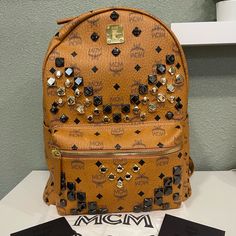 This Is A Cognac Medium Stark Visetos Studded Logo Backpack. Everything You Could Want In A Fashionable Pack Is Included In This Logo-Stamped Bag Peppered With Gleaming Pyramid Studs: A Padded Pocket For Safekeeping Your Ipad Or Other Tech Gear; A Roomy Interior And Exterior Zip And Slip Pockets For Stowing Sunglasses And Other Small Essentials. 12 1/2"W X 16"H X 6 1/2"D. 3.65 Lbs. Dual Top-Zip Closure Top Handle; Adjustable Backpack Straps Exterior Zip And Slip Pockets Interior Zip, Wall And Sm Designer Backpack With Detachable Strap For Shopping, Luxury Leather Shopping Backpack, Luxury Leather Backpack For Shopping, Designer Standard Backpack With Branded Hardware, Luxury Standard Backpack With Branded Hardware, Designer Leather Backpack With Branded Hardware, Luxury Leather Evening Backpack, Mcm Bags, Tech Gear