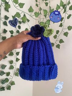 a hand holding a blue knitted beanie with a flower on the top and green leaves around it