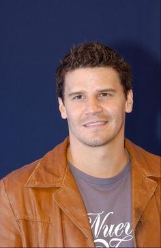 a man wearing a brown leather jacket smiles at the camera while standing in front of a blue wall