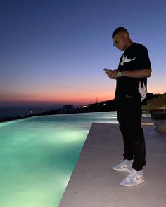 a man standing on the edge of a swimming pool looking at his cell phone as the sun sets