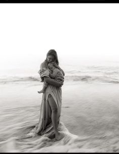 a woman holding a baby in her arms while standing on top of a beach next to the ocean