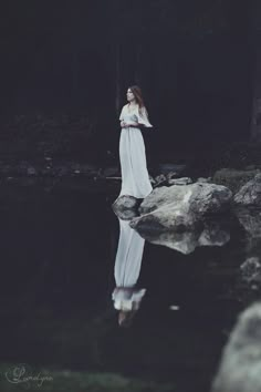 a woman in white dress standing on rocks near water with trees and rocks behind her