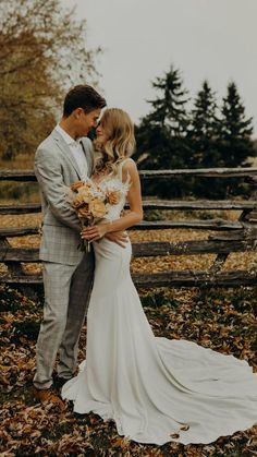 a bride and groom kissing in front of a wooden fence with autumn leaves on the ground