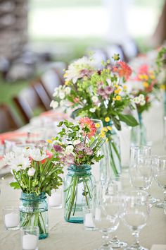 several vases filled with flowers on top of a long table covered in wine glasses