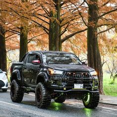 a black truck is driving down the road in front of some trees with orange leaves