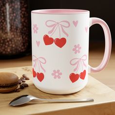 a pink and white coffee mug with hearts on it next to a spoon, cookie