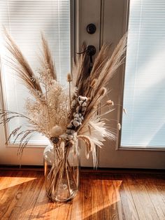 dried flowers in a glass vase sitting on the floor next to a door with blinds